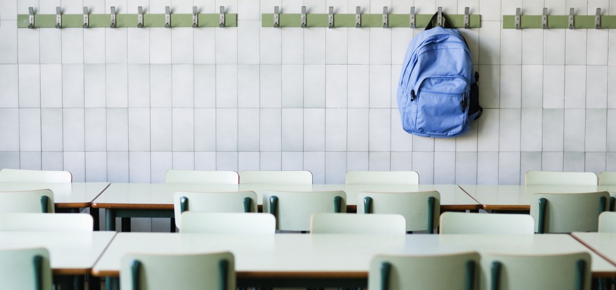 A row of desks in a classroom with a backpack hanging up on the back wall