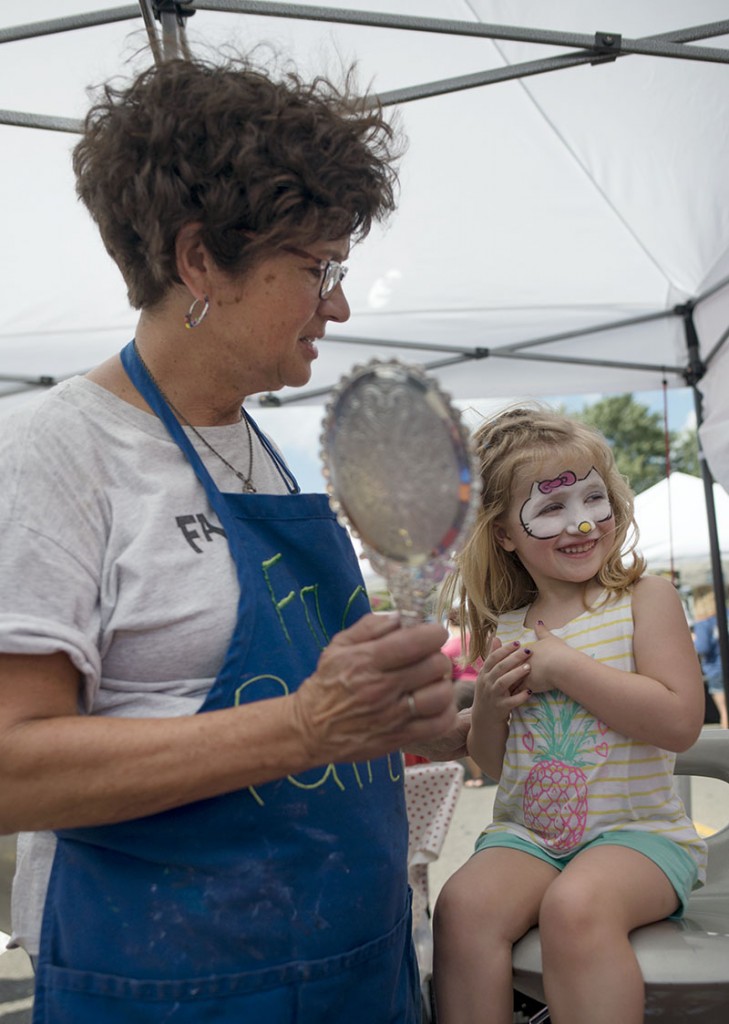 Susie Kennedy, left, holds out a mirror to show Adalynne Schwartz, 4, her Hello Kitty face painting while she grins at her dad at the Lithopolis Honey Bee Festival in Lithopolis, Ohio on Saturday, Sept., 10, 2016.  For $500, Face Splash Face Painting booth would transform kids into their favorite characters, like Elsa from Frozen and Pikachu. (Kelsey Brunner/WOUB)