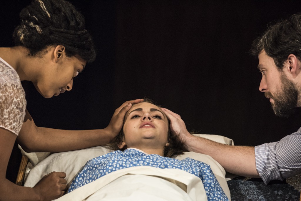The opening scene of "The Library," in which Caitlin Gabriel (middle), played by Rachel Gaunce, appears on stage in a hospital bed. She is injured from what the audience learns was a school shooting and is surrounded by her father Nolan Gabriel (right), played by Carson Cerney, and her mother Elizabeth Gabriel, played by Sana Selemon. (Robert McGraw/WOUB)