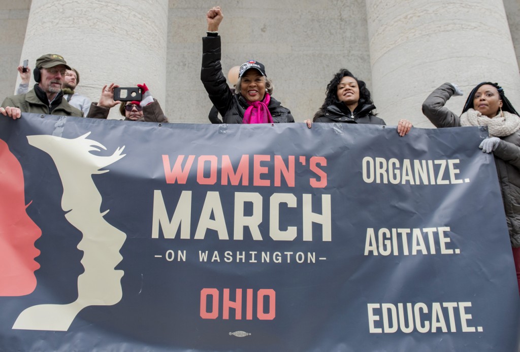 Wearing a pink scarf, U.S. Congresswoman Joyce Beatty 3rd District of Ohio stands on the Statehouse steps with Columbus City Councilmember, Jaiza Page, and Ohio Women’s March Co-Organizer, Rhiannon Childs, as they hold the rally banner on Sunday afternoon, January 15, 2017. (Margo Sabec/WOUB)