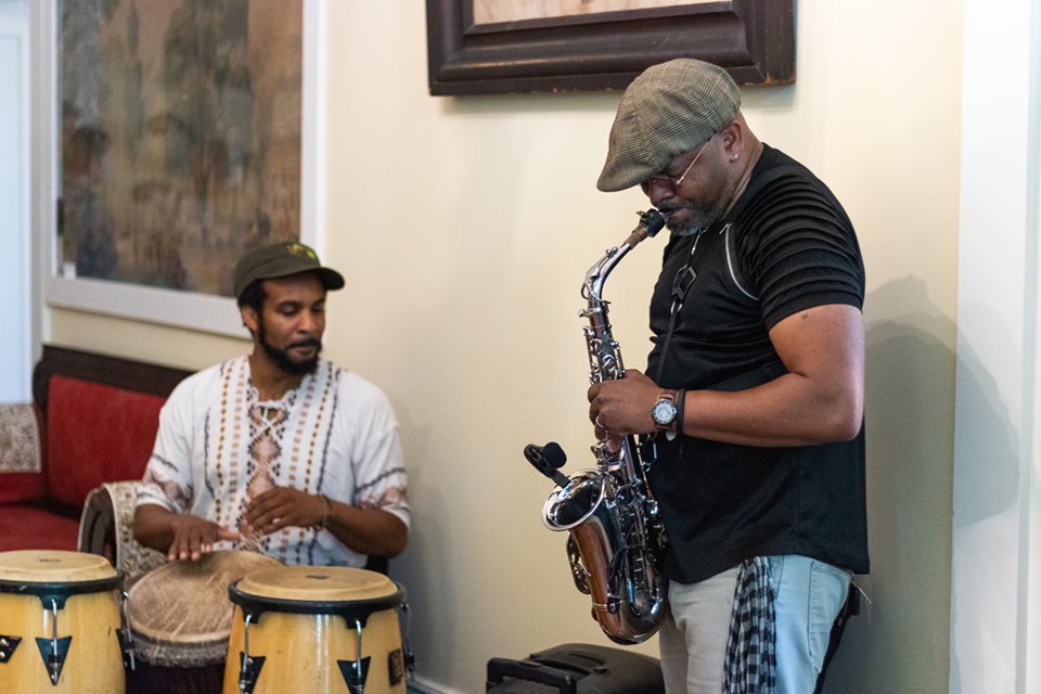 A man plays the sax while another plays hand drums
