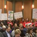 Protestors stand in crowded meeting with signs.