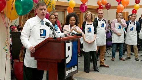 man at podium, six others nearby all wearing Goodwill aprons