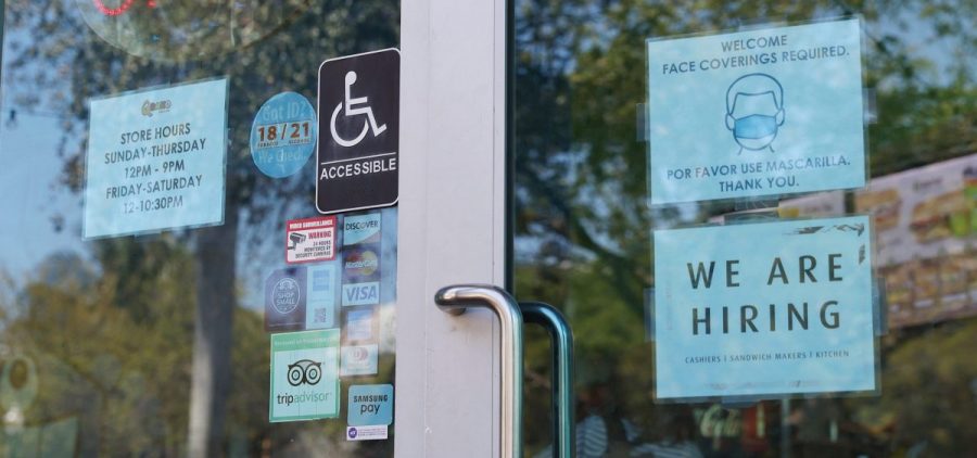 A "We are hiring" sign is paired with a pandemic-themed sign in the window of a store in Miami. Restaurants and other in-person businesses are looking to hire more workers at a time when some are wary of returning to work or are busy with caregiving.