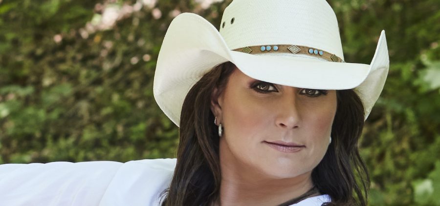 Terri Clark poses for a promotional photo in a wooded area. Terri Clark is wearing a white cowboy hat and white blouse.