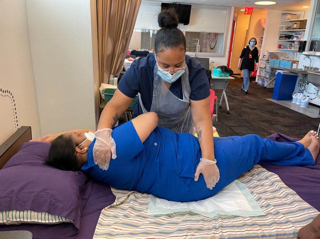 Juhaley Abrau, a student at Cooperative Home Care Associates, practises how to safely give her fellow student, Tashawna Vivas, a bedbath. Abrau says a lot of the programs she looked at were all "insanely expensive," in some instances, charging up to $2,000. She chose this program because it was free, paid for through government and private money.