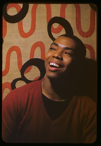 Portrait of Alvin Ailey red v-neck sweater smiling, against burlap backdrop with red/black squiggles.