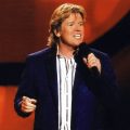 Peter Noone of Herman's Hermits in a promotional photo that features an image of Noone on stage holding a microphone against a red backdrop.