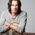 Rick Springfield poses with a guitar against a gray background for a promotional photo.