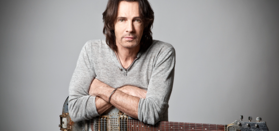 Rick Springfield poses with a guitar against a gray background for a promotional photo.