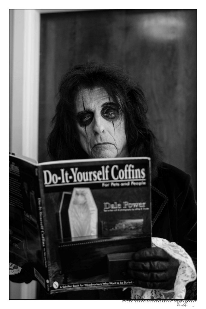 Alice Cooper poses for a promotional photo with a book entitled "Do-It-Yourself Coffins."