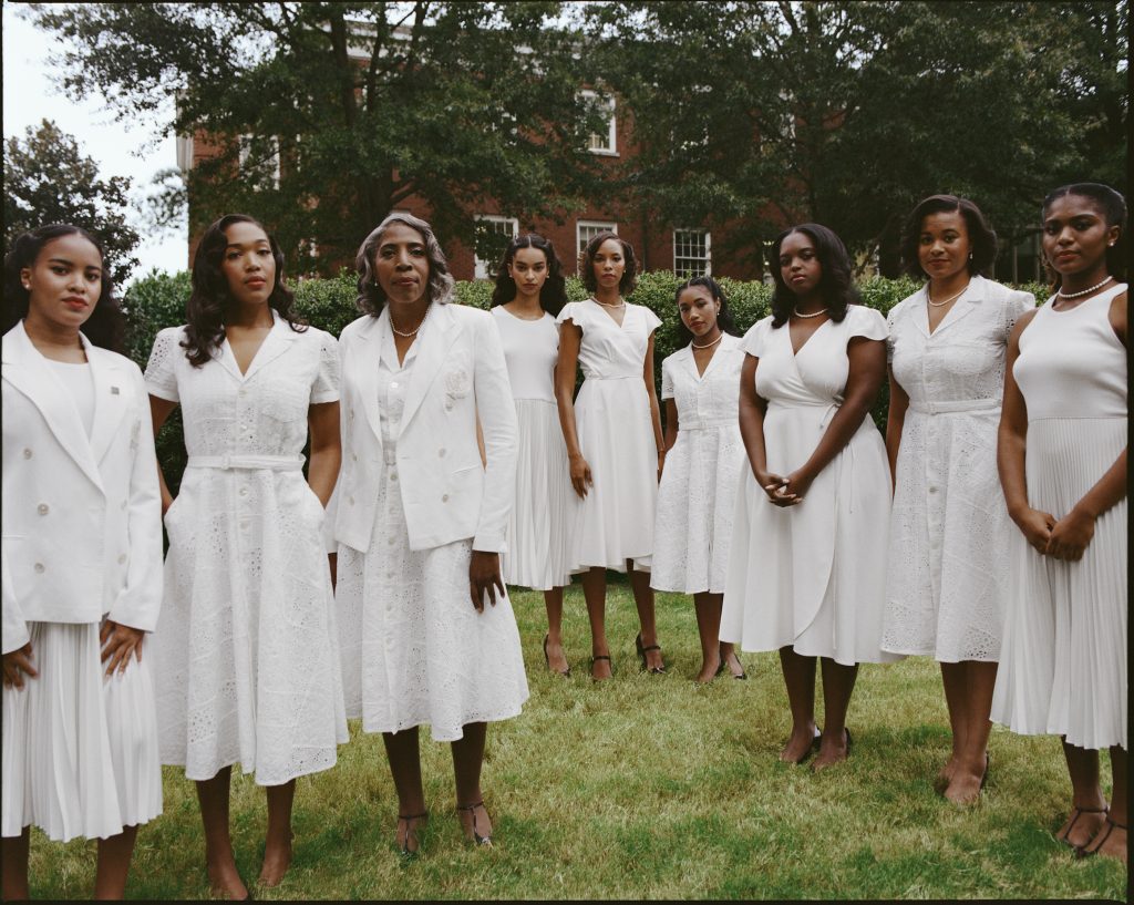 Nine Black women of various ages stand shoulder to shoulder on a college campus in white dresses
