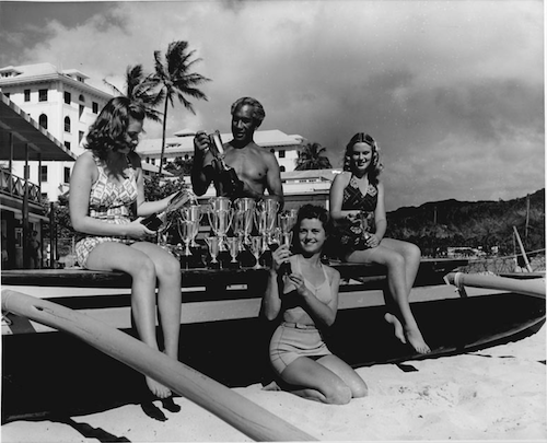 man standing behind surfboard with many trophies. Three women in swimsuits with him smiling