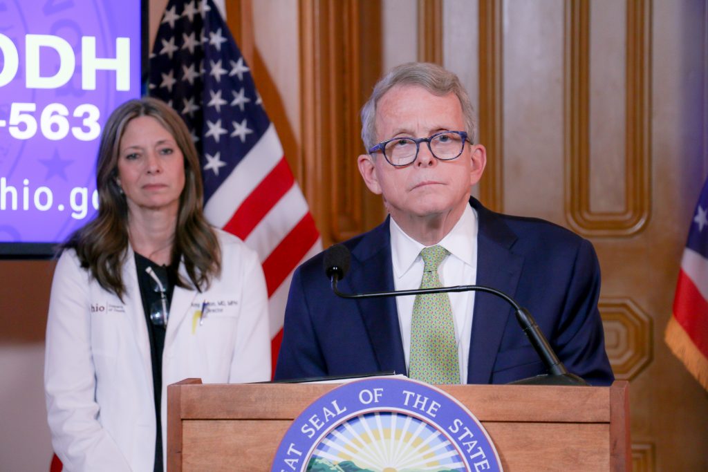Gov. Mike DeWine stands at a podium during a press briefing with Dr. Amy Acton over his shoulder