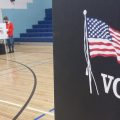 A person votes off in the distance. They are in a school gym. A box in the foreground has an American flag on it and the word Vote underneath it.