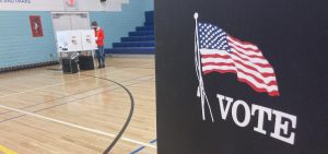 A person votes off in the distance. They are in a school gym. A box in the foreground has an American flag on it and the word Vote underneath it.