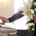 A person with flowers in their hand lays their other hand on a casket at a funeral