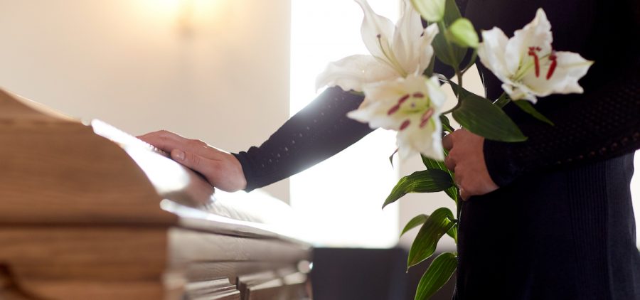 A person with flowers in their hand lays their other hand on a casket at a funeral