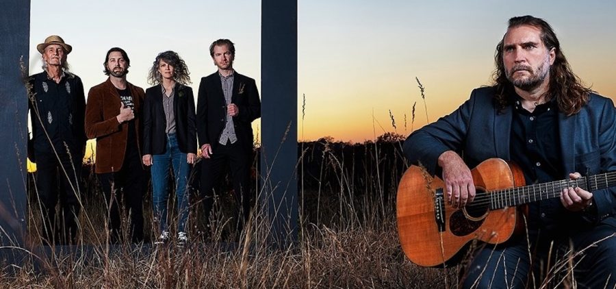 A promotional picture for the band June Star. The band poses in a field at dusk. Lead singer and chief songwriter Andrew Grimm sits in the foreground with an acoustic guitar, and additional band members stand within a frame in the distance behind Grimm..
