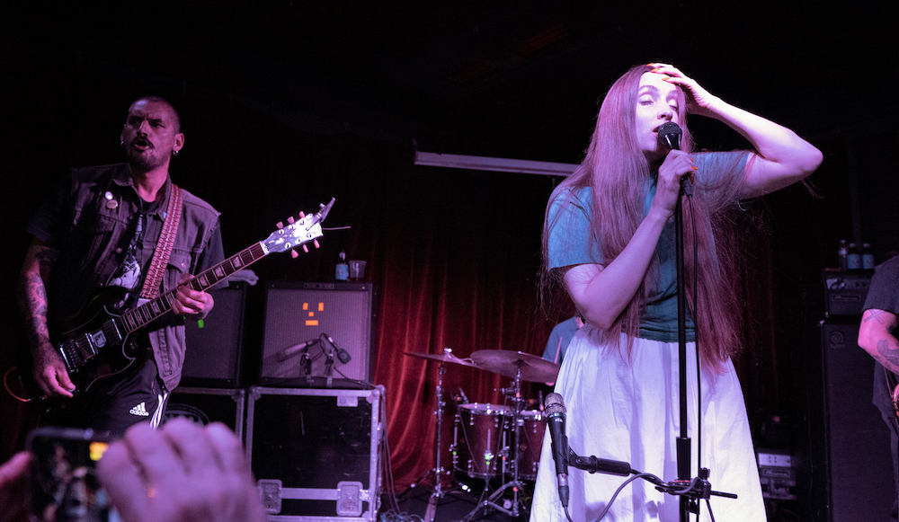 Dry Cleaning performs at Ace of Cups in Columbus, OH. Pictured from left to right: guitarist Tom Dowse, vocalist Florence Shaw. Shaw is emoting dramatically, with one hand on her head and one clutching the microphone.