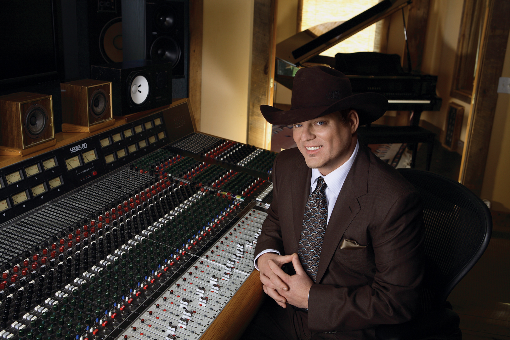 A promotional image for musician John Michael Montgomery. Montgomery is in a darkened recording studio, leaning on a mixing board and wearing a cowboy hat.