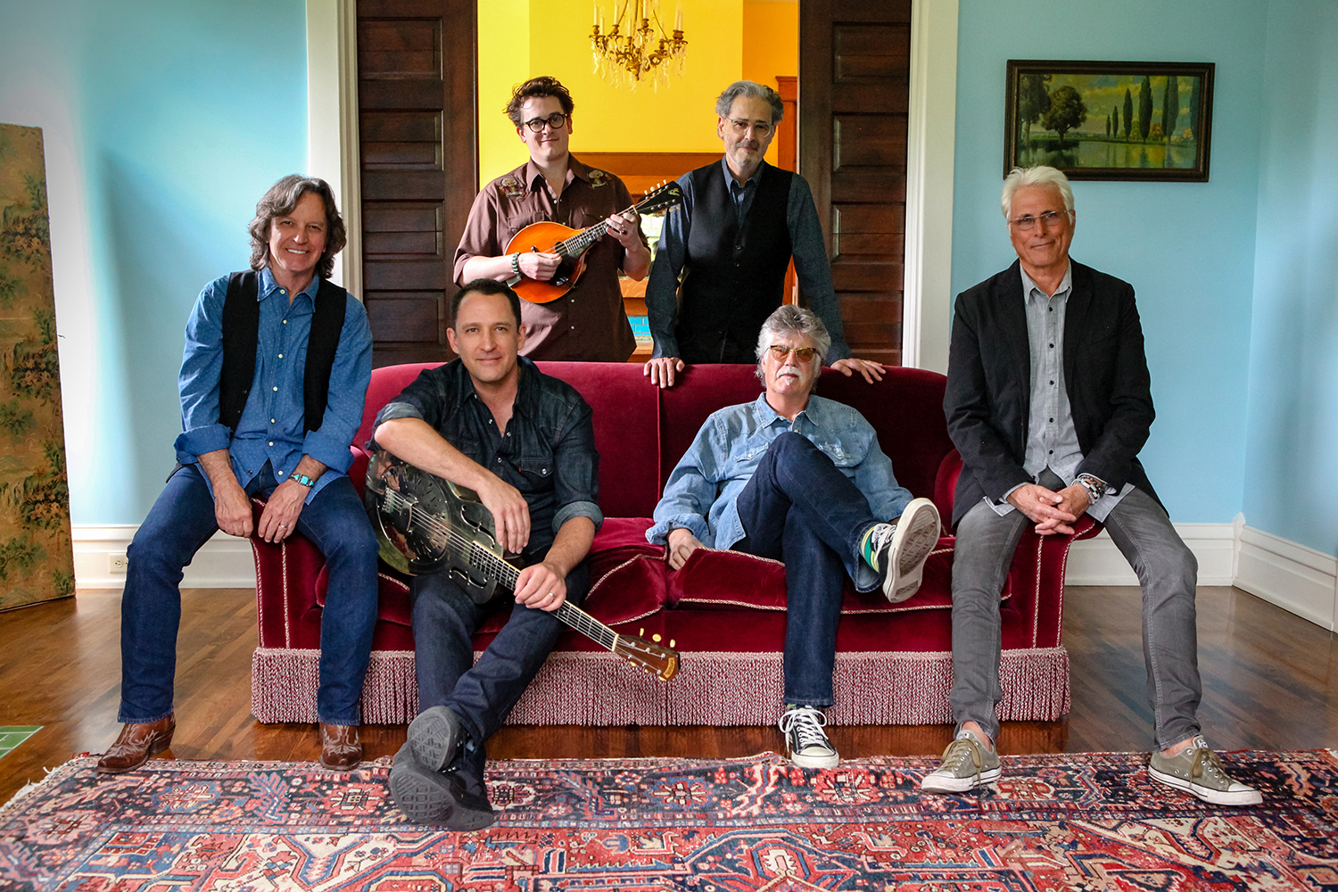 A promotional photo of the Nitty Gritty Dirt Band. Four of the six band members are sitting on a red couch, and the two remaining members stand behind them.
