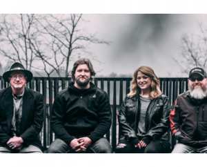 A promotional image of the Sour Mash String Band, the band is sitting on a bench against a cloudy sky.