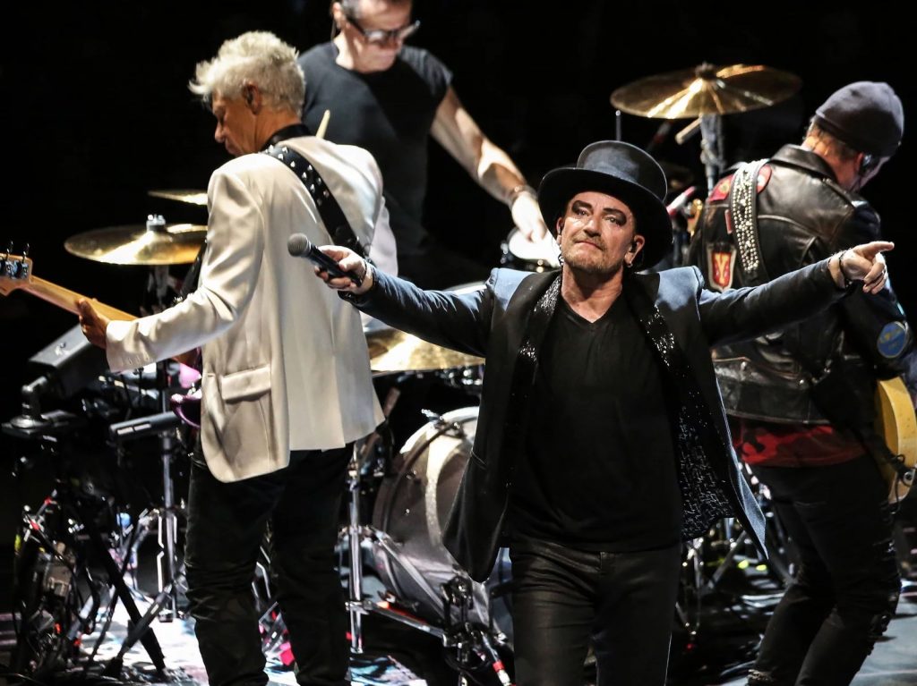 The irish band U2 during a 2011 performance in Paris. Pictured are Irish band U2's bass Adam Clayton, drummer Larry Mullen Junior, singer Bono and guitarist The Edge. Bono is in the foreground with heavy black eye makeup, a top hat, and arms outstretched. The rest of the band faces away from the camera.