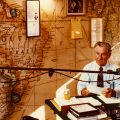 Lowell Thomas at desk with world map behind him as wallpaper