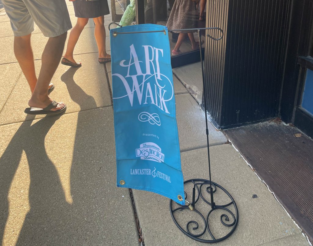 A blue flag that reads "Art Walk" hangs on a small shepherd's hook in the streets of Lancaster during the 2022 Lancaster Festival Art Walk.