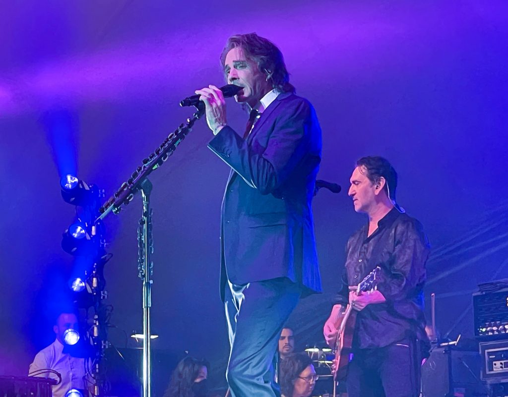Rick Springfield holds a microphone while performing at the Lancaster Festival. The stage has a purple and blue color scheme and Rick Springfield is wearing a suit.