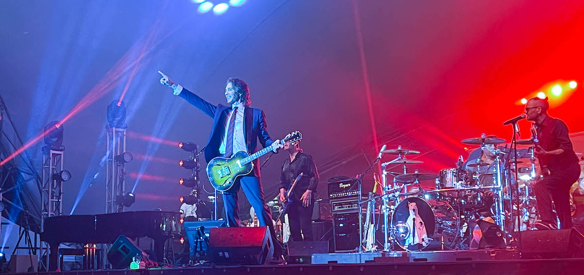 Rick Springfield points upward and outward to the enthusiastic crowd during his performance at the 2022 Lancaster Festival. Rick Springfield is on stage and is wearing a suit and holding a guitar. His band is set up in the background.