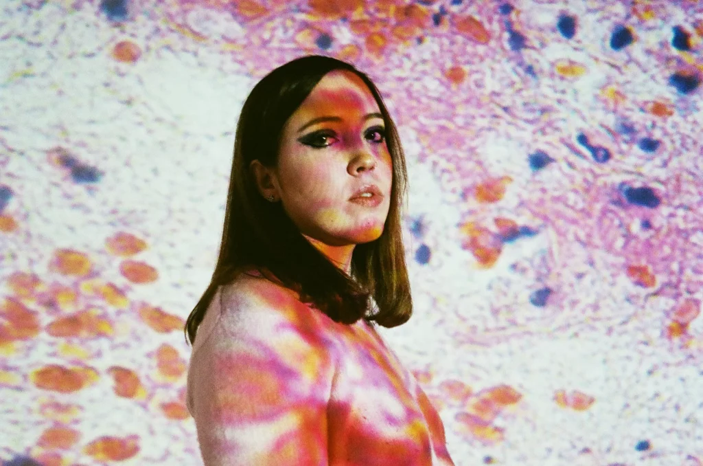 A promotional photo of Sophie Allison, who performs as Soccer Mommy. Allison is posing against a white background with a colorful, psychedelic pattern superimposed over her.