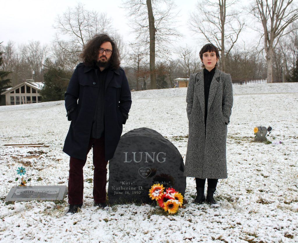 A promotional picture for the band Lung. Members Daisy Caplan and Kate Wakefield are standing in a snowy cemetary with barren trees behind them, in front of a headstone that reads: LUNG.