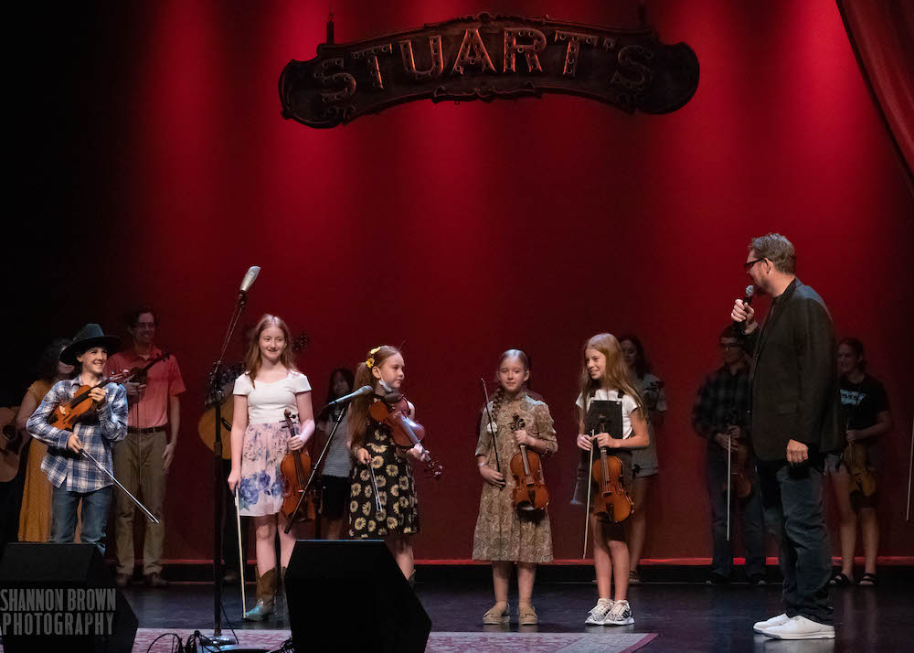 The youngest competitors in the Ohio State Old Time Fiddlers' Contest stand on the stage at Stuart's Opera House. Five children are standing in the foreground in front of microphones, fiddles in hand, while an adult judge speaks into the microphone.