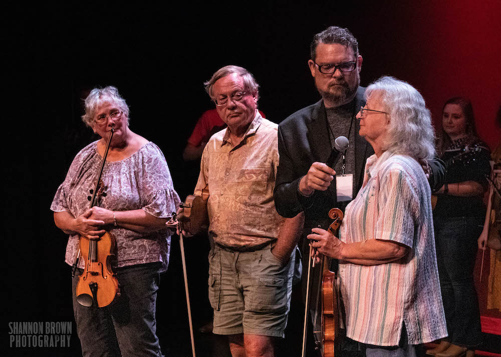 Four people are on the stage at Stuart's Opera House. They are lined up on the stage, and everyone is holding a fiddle. Two of the people are female presenting, and two of them are male-presenting. Three of the four are holding fiddles and the last one is holding a microphone.