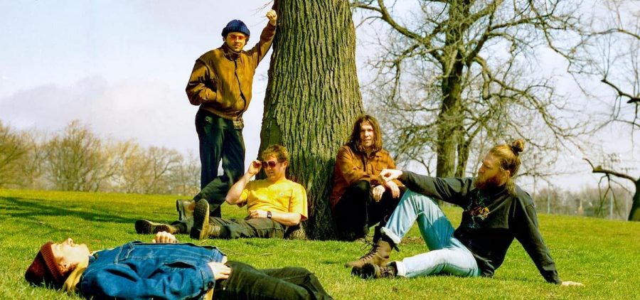 A promotional image for the band In the Pines. Four male-presenting people with light skin are wearing winter clothing on a bright day in a field underneath a barren looking tree.