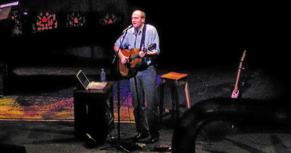 James Taylor performs in the spotlight on a stage, holding a guitar with amps behind him. 