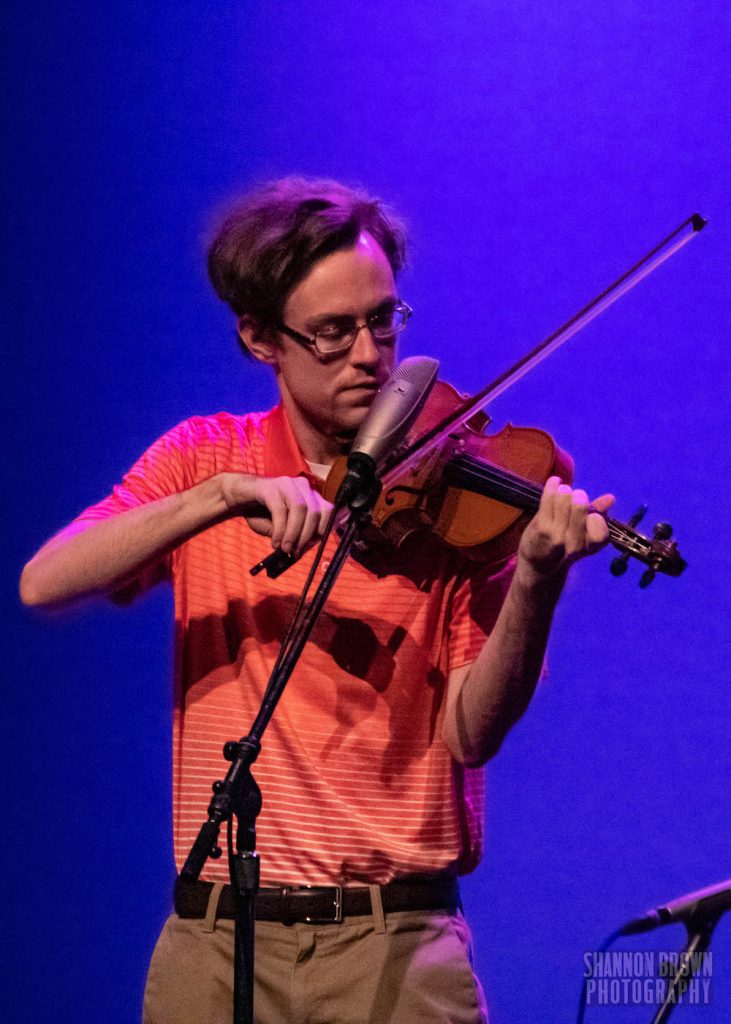 A male-presenting person with short, brown hair is playing a fiddle intently on a stage with a purple backlight. They are wearing an orange short sleeved shirt and brown slacks. 