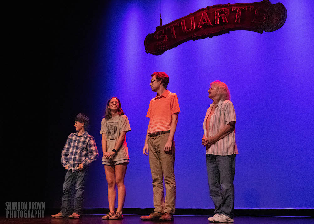 Four people stand on the stage of Stuart's Opera House. There is one male presenting very young child, a slightly older female-presenting child, a tall, male presenting adult, and a slightly shorter female-presenting person.