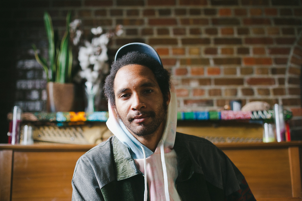 A promotional headshot for artist Tre Burt. Burt has dark skin and is wearing a hoodie with a jacket over it, posing against a bare brick wall with a short, out of focus table behind him. 