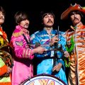 Four male-presenting people in the same marching band inspired costumes The Beatles wore on the cover of "Sgt. Pepper's Lonely Hearts Club Band." From left, the person is in a lime green suit with red trim, like John Lennon, the next person in a pink suit with red trim, like Ringo, the next person in a blue suit with dark blue trim like Paul McCartney, and the last person in a yellow suit with red trim and a big funny hat, like George Harrison.