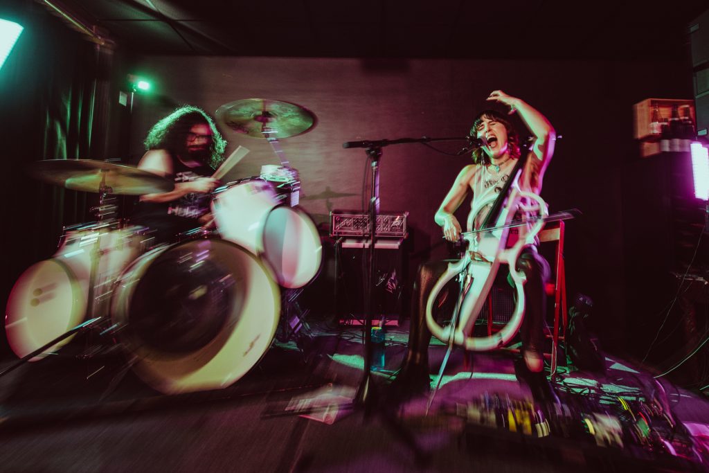 The band LUNG plays a dark rock club. This is a promotional picture, and drummer Daisy Caplan is drumming while Kate Wakefield is singing into a microphone and playing cello. 