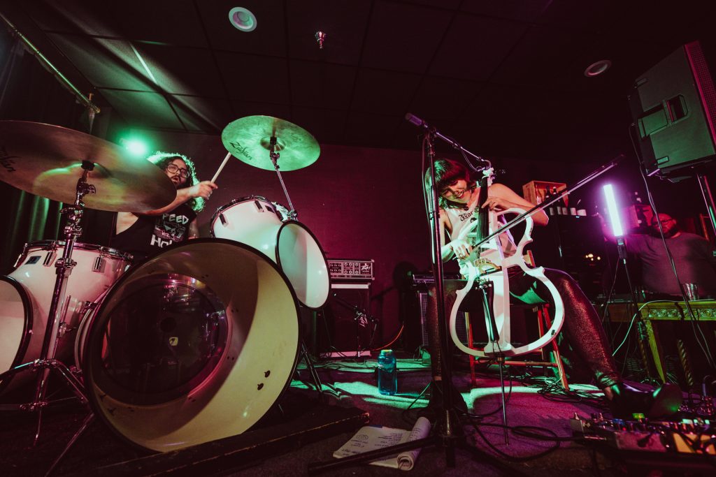 A promotional picture for the band Lung. Drummer Daisy Caplan is drumming and Kate Wakefield is singing and playing the cello. The band is in a darkened rock club.