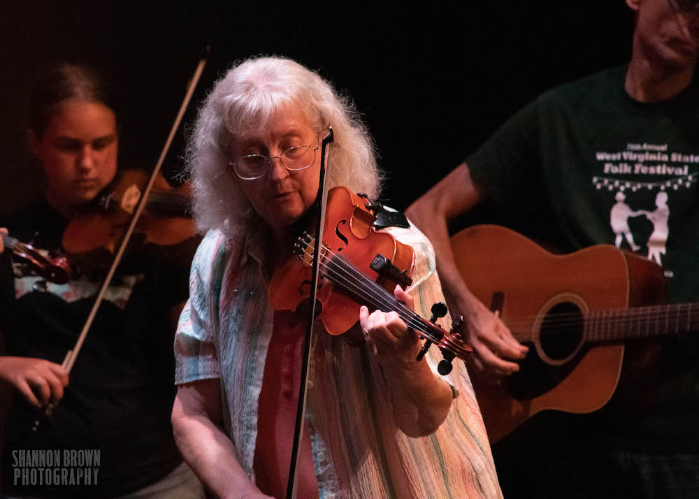 A female-presenting person with white, shoulder length hair and bangs is playing the fiddle. They are also wearing glasses.