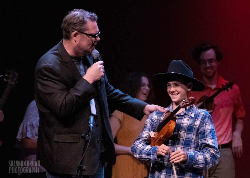 A young male presenting person with a cowboy hat and a plaid shirt on smiles while clutching a fiddle as they are named the winner of the age 12 and under category. 