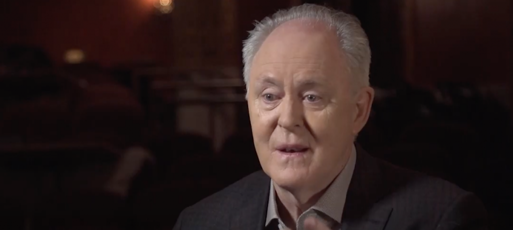 John Lithgow is pictured as a talking head for the PBS program "On Broadway." Lithgow is wearing a brown suit in a darkened room.