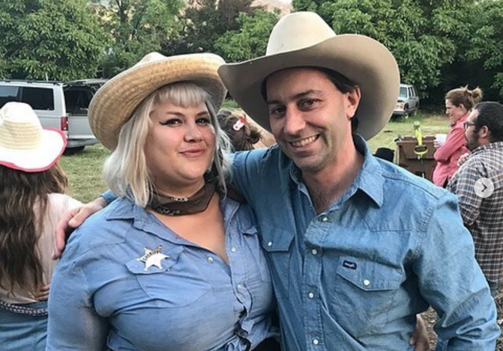 Two people dressed in denim shirts and cowboy hats lean in, smiling, for a picture. On the left hand side is a female presenting person, Shannon Shaw. On the right is a male-presenting person is Joe Haener. They are outside on a sunny day.