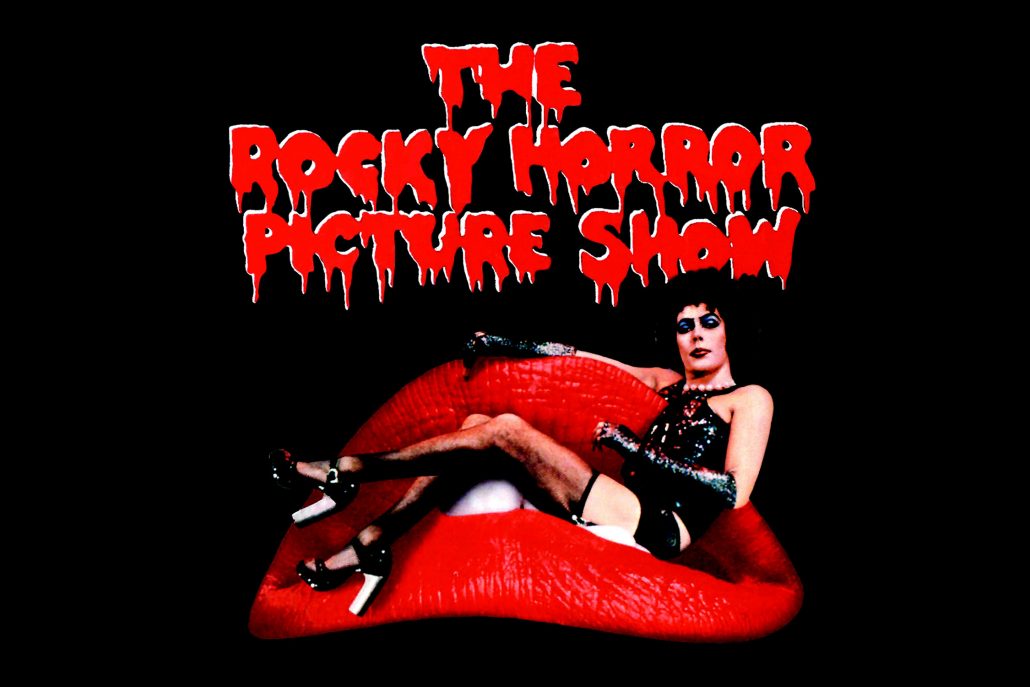 A promotional image for "The Rocky Horror Picture Show." The image features someone wearing a shiny black outfit and platform shoes sitting in an enormous pair of red lips.