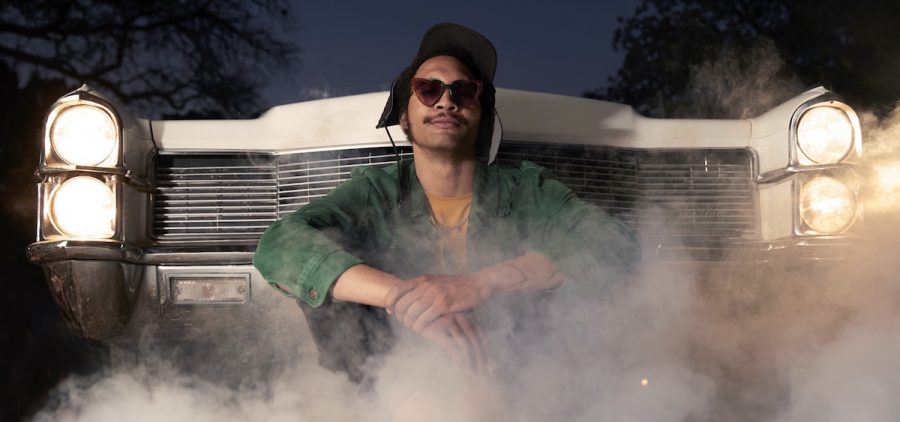 A promotional image for artist Tré Burt. Burt is pictured sitting in front of a car, the car is smoking and Burt is wearing sunglasses, a jacket, and a hat.